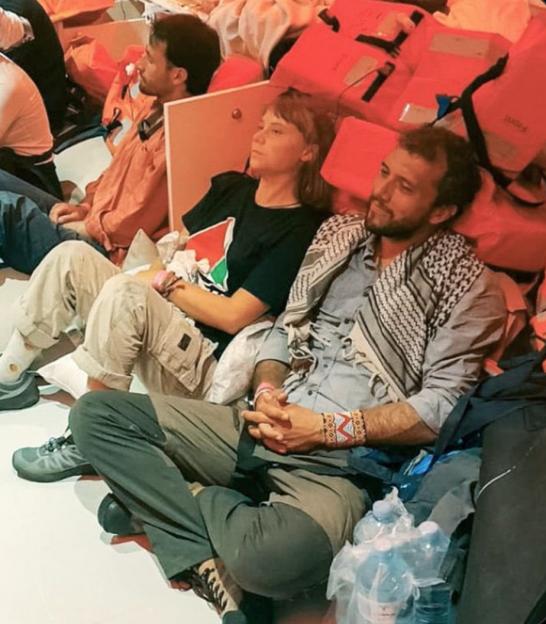 People sitting on the floor, surrounded by life vests and bottled water, with one woman wearing a "Free Palestine" T-shirt and one man wearing a keffiyeh.
