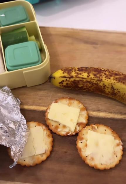 Crackers with butter or cheese, a banana, and a segmented lunchbox on a wooden board.