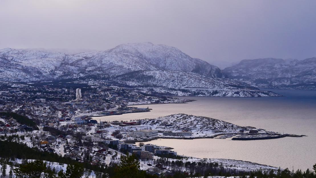 Panoramic view of the snow-covered city of Alta, Norway, featuring Altafjord and mountains on a cloudy winter day.