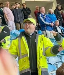 A man in a bright yellow safety jacket leads a crowd in a stadium.