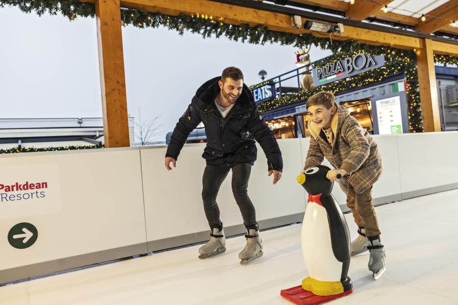 A man and a boy ice skating, with the boy using a penguin-shaped ice skating aid.