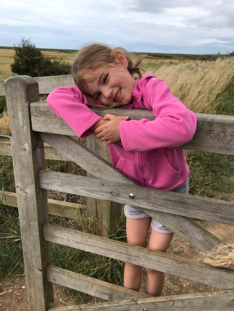 Emily, a young girl, smiles while leaning on a wooden fence outdoors.