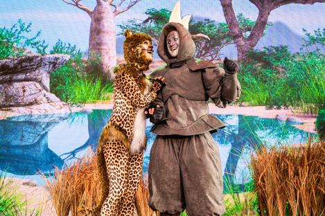 Two performers in cheetah and rhinoceros costumes on a stage with a savanna backdrop.