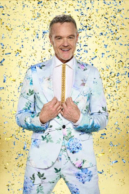 Stefan Dennis wearing a light blue floral suit, white shirt, and golden beaded tie against a yellow background with falling blue and gold confetti.