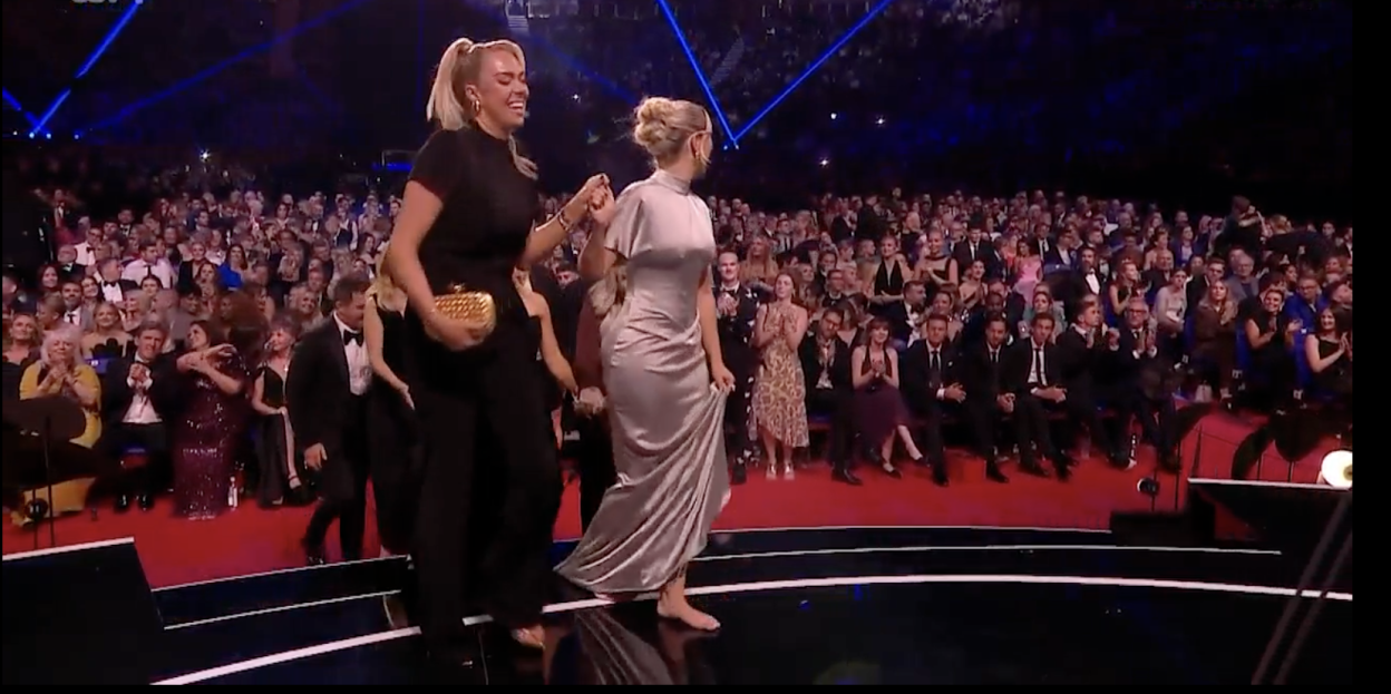 Two women walking barefoot across a stage to applause.