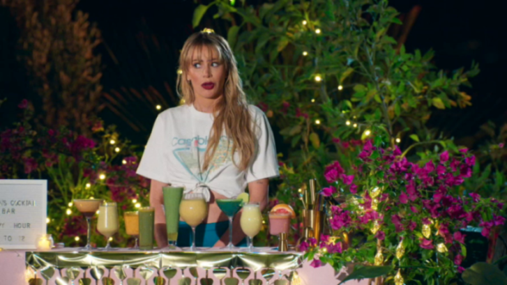 A woman with blonde hair, wearing a white t-shirt, stands behind a bar with several colorful cocktails.