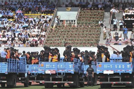 People in custody being transported in trucks, surrounded by police and observed by spectators in a stadium.