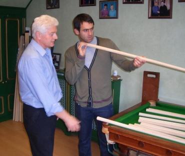 Two men examining a long cue stick next to a pool table.