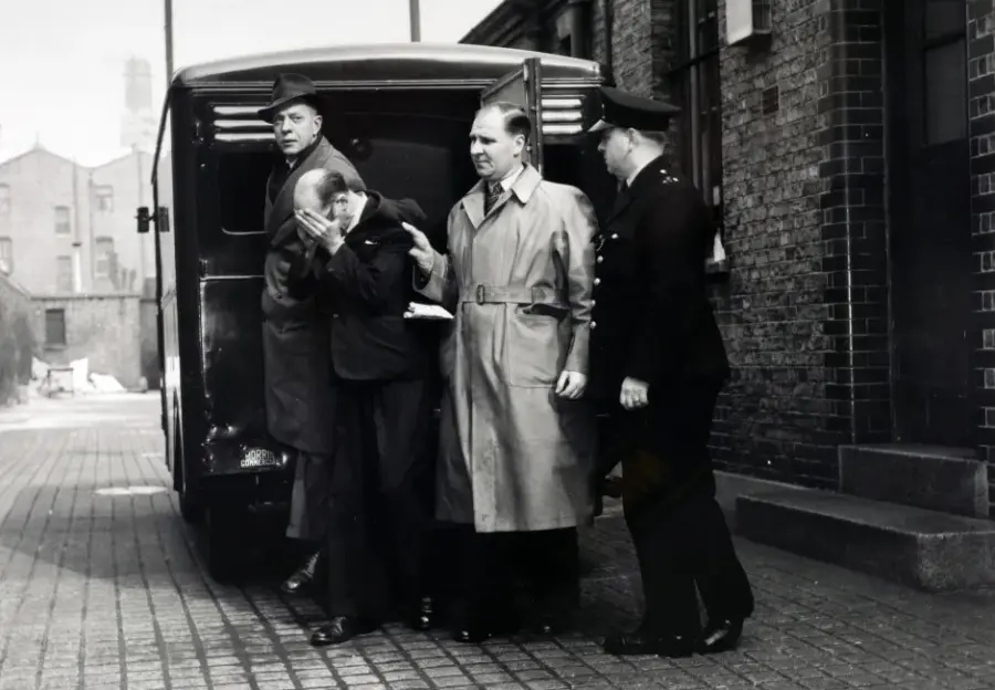 Black and white photo of a man in custody being escorted by police officers.