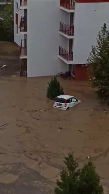 A flooded street in Elenite, Bulgaria, with a partially submerged white car and an apartment building in the background.