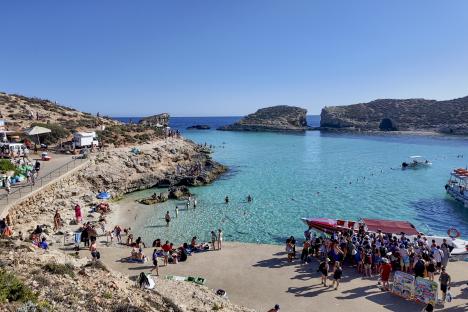 Aerial view of Blue Lagoon, Camino, Malta, showing many people swimming and relaxing on the beach and rocky coastline.
