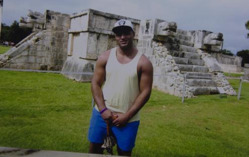 Man in tank top and shorts standing in front of Mayan ruins.