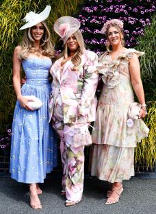 Three women in elegant outfits and hats pose in front of a floral wall at a racecourse.
