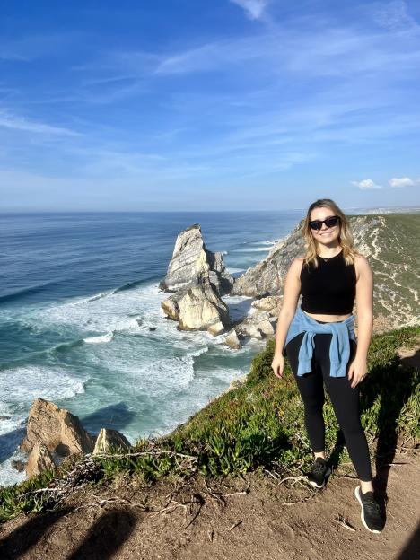 A woman is standing on the edge of a cliff, looking out at a beautiful ocean view.