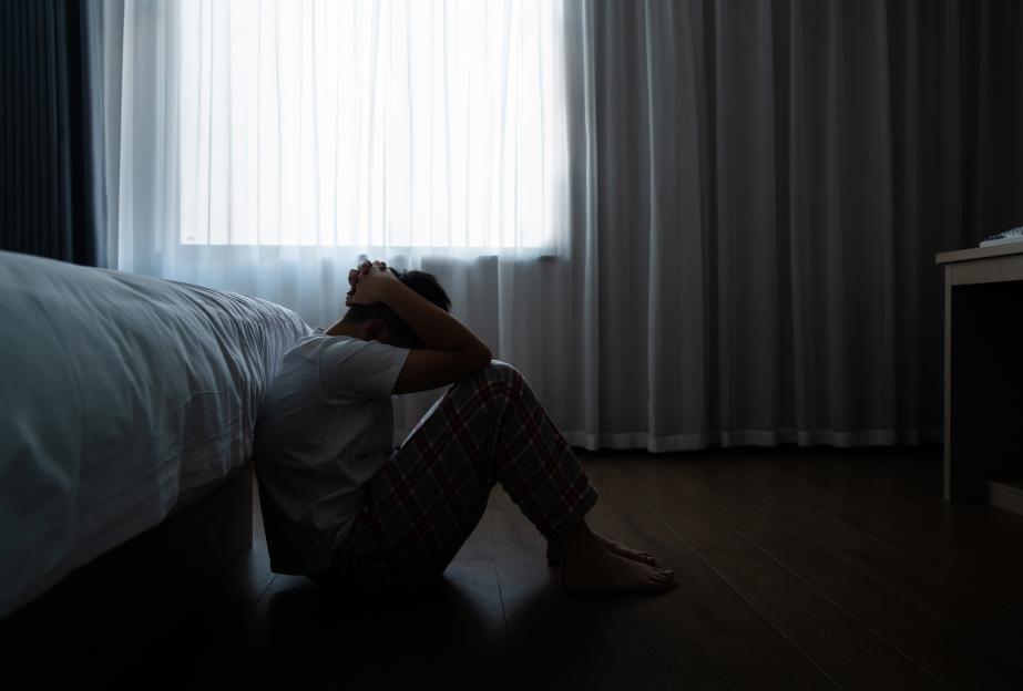 A depressed teenage boy sits on his bedroom floor, head in hands.