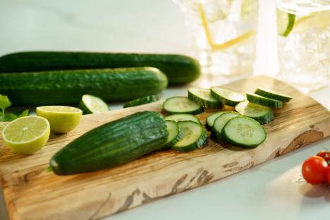 Sliced cucumbers and limes on a wooden cutting board next to lemonade.