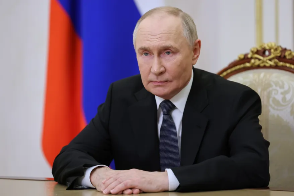 Russian President Vladimir Putin sitting at a table with his hands clasped, in front of the Russian flag.
