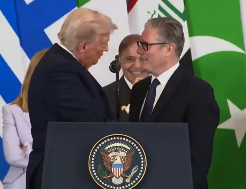 Donald Trump speaking at a podium with the US presidential seal, turning to a smiling man with flags behind him.