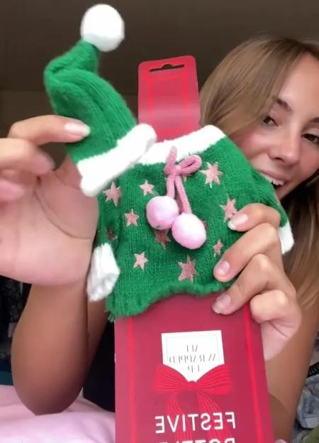 A person holding up a green Christmas elf outfit with pink stars and pom-poms on a red cardboard hanger.