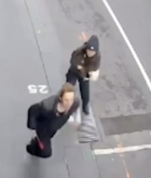 High-angle view of two people on a street, one in a dark hoodie appears to be pushing the other, who is dressed in black and looking up at the camera.