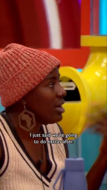A woman in a red beanie and vest speaks, wearing earrings shaped like the continent of Africa.