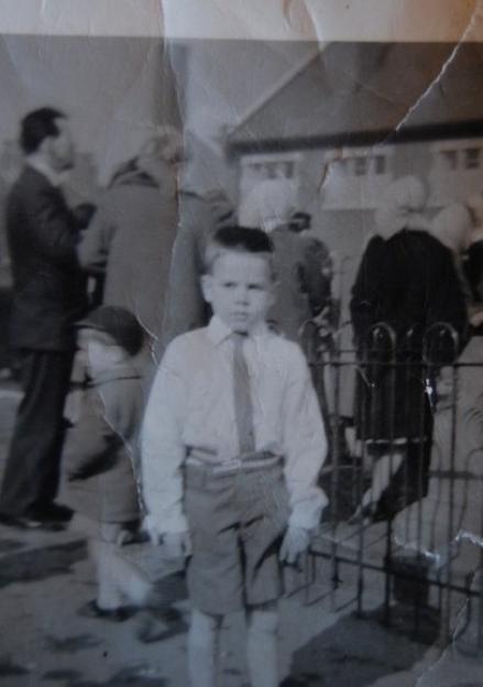 Tommy Rhattigan as a young boy, dressed in a shirt and tie, standing in a street.