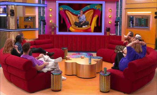 JoJo Siwa on a large screen, watched by housemates on a red sofa.