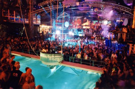 A crowded nightclub with a pool and a DJ booth.