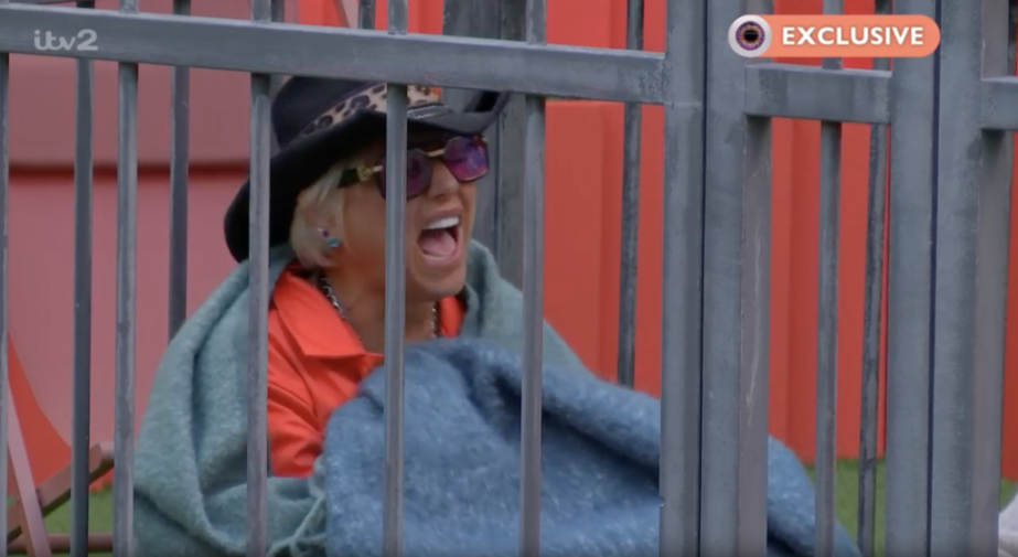 A person with a blonde bob, wearing sunglasses, a black cowboy hat with leopard print, an orange shirt, and a blue blanket, is laughing with their mouth wide open behind gray bars.