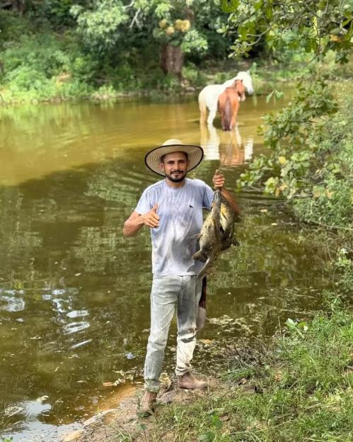 Reinaldo da Silva Melo, an influencer known as "King of Juquira", holding caught fish, giving a thumbs-up, with two horses behind him in a river.