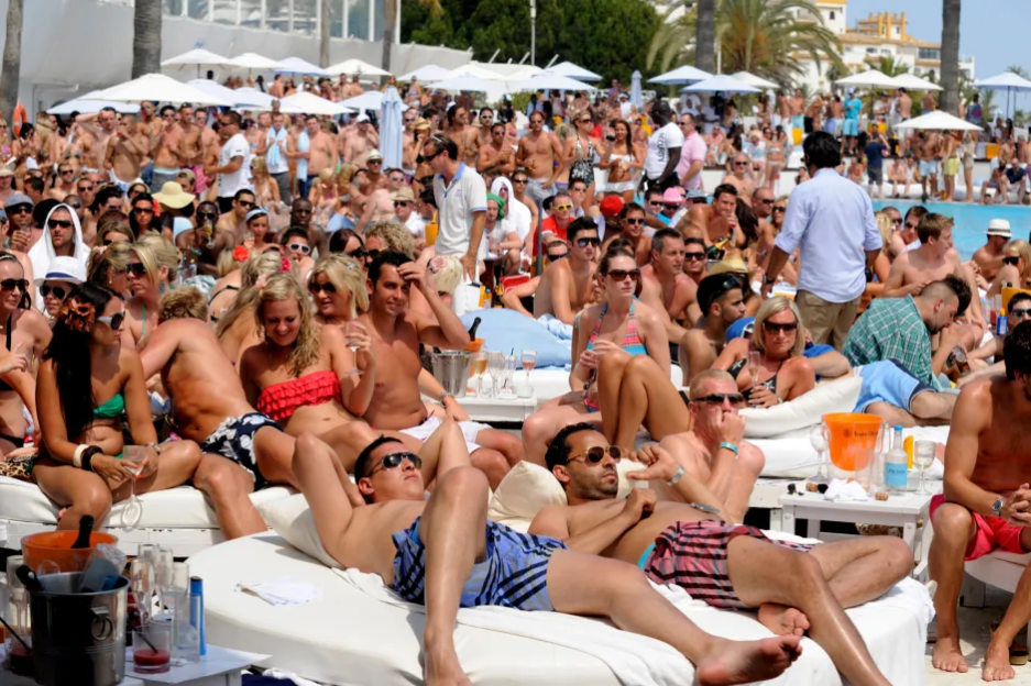 Crowded beach club scene with people lounging on white daybeds.