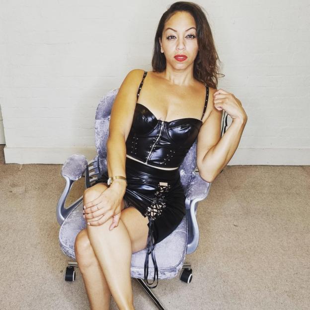 Zara the dominatrix in a black leather outfit, seated in a chair.