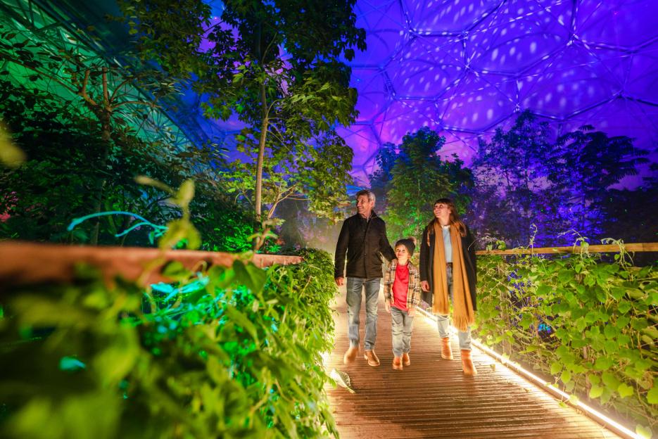 A man, woman, and child walking in the Eden Project dome, lit by green and purple lights.