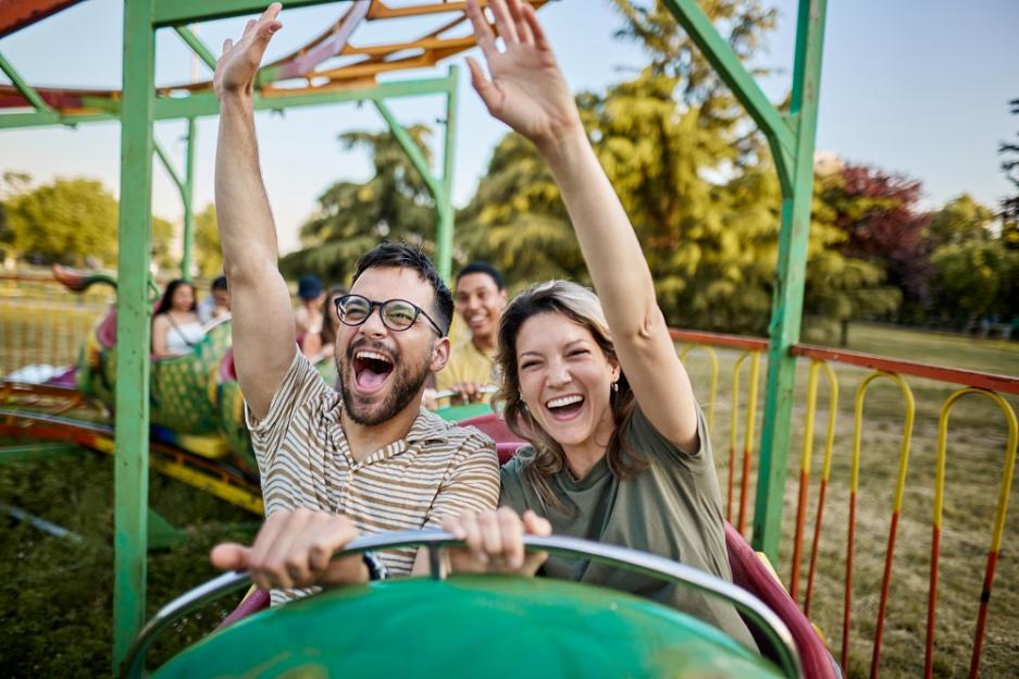 Happy couple riding on rollercoaster with raised arms at amusement park.