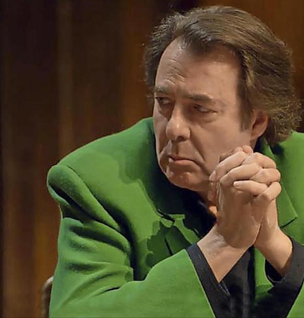 Man in a green jacket with his hands clasped, looking thoughtfully to the left.