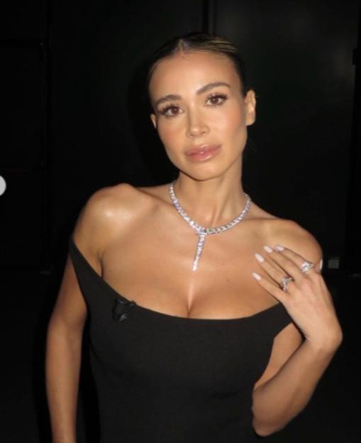 A woman with light brown hair, pulled back, wears a black off-the-shoulder dress, a sparkling necklace, and rings, while posing against a dark background.