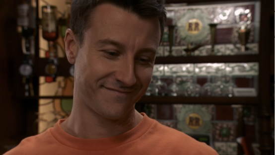 Man in orange shirt smiling subtly.