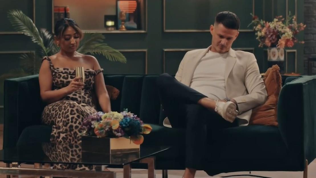 Steven and Nelly sitting on a dark green couch with a coffee table with colorful flowers in front of them, Steven on the right with his leg crossed, Nelly on the left holding a glass of champagne.