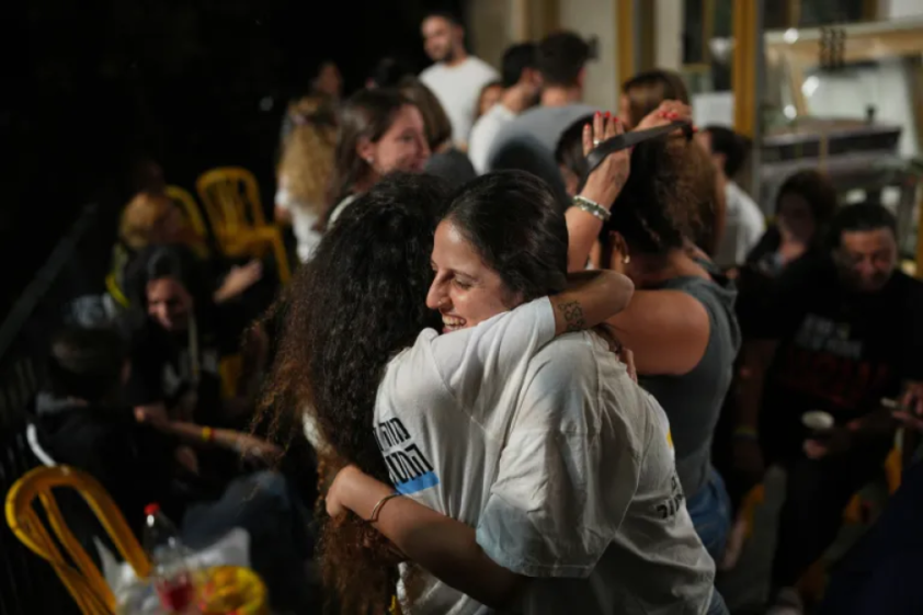 Two women embrace, smiling, while surrounded by a crowd of people.