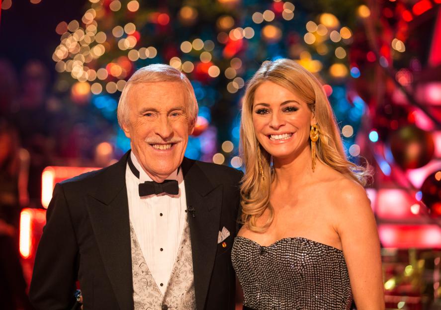 Sir Bruce Forsyth and Tess Daly smiling, dressed in formal wear, in front of blurred lights.
