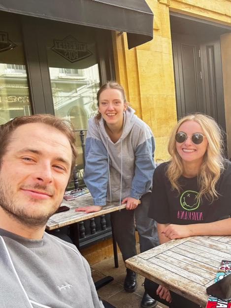 Max Bowden taking a selfie with two women, Katie McGlynn in sunglasses, sitting at outdoor tables.
