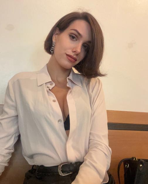 Noelle posing in a light pink button-up shirt with an unbuttoned top revealing a black bra.