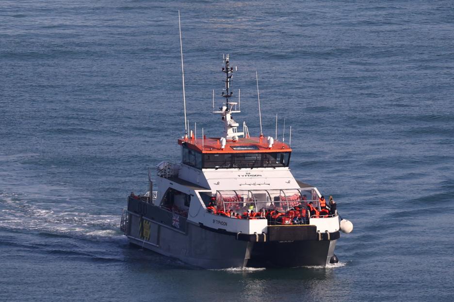 A Border Force vessel, the BF Typhoon, carries migrants across the sea, with many people visible on deck wearing orange life vests.