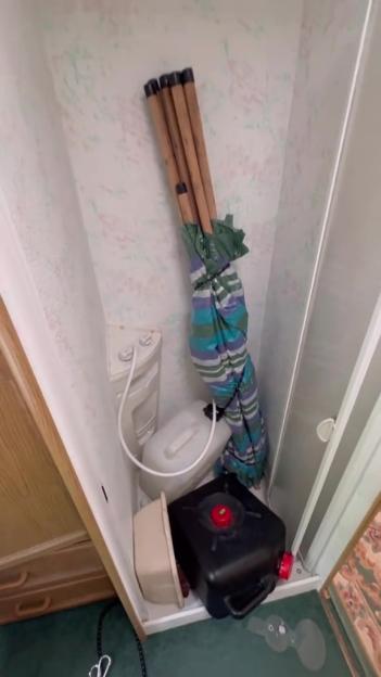 A narrow closet in a caravan with a small white sink, two plastic containers on the floor, and a colorful umbrella leaning against the wall.