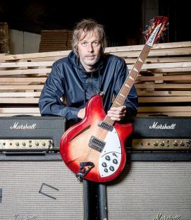 A man holding a red Rickenbacker guitar in front of two Marshall amplifiers.