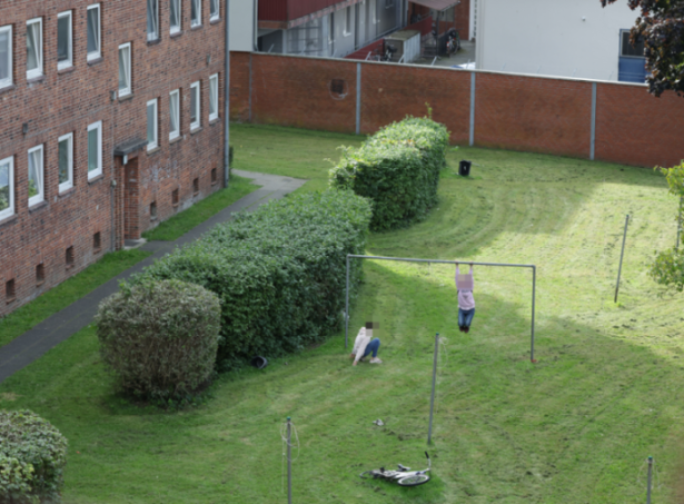 Two people in a yard, one hanging from a metal bar with a box over their head, the other sitting on the grass.