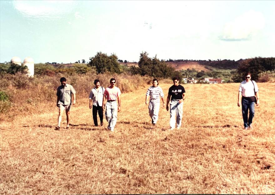 Six men in casual clothing walk across a dry, grassy field.