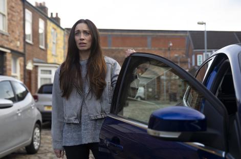 A worried woman stands by a car on a residential street.