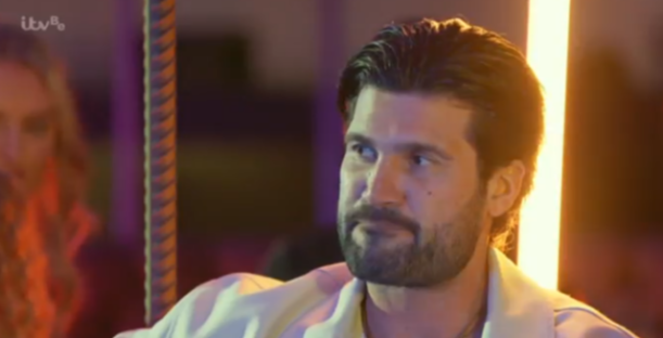Screenshot of a man with dark hair and a beard, looking pensive.