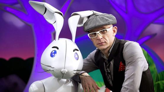 A man with a newsboy cap and glasses poses with a robotic rabbit.